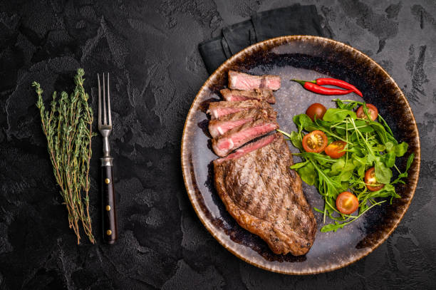 istock Grilled striploin steak, beef meat steak on a plate with salad. black background. top view 2198404638