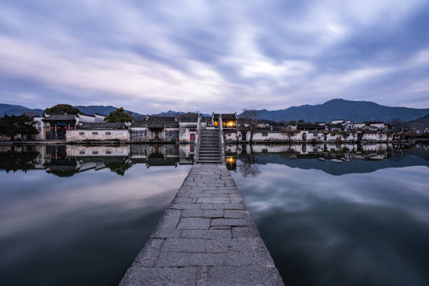 This mesmerizing photograph captures a tranquil dawn scene in a historic Chinese village, where a stone pathway leads the eye towards a gracefully arched bridge and traditional white-walled houses reflecting in the still waters of a pond. The long-exposure effect gives the sky a soft, flowing texture, enhancing the serene and timeless atmosphere of the setting.\n\nWarm lights glowing from within the village contrast with the cool blue tones of the twilight sky and water, adding a sense of warmth and inviting nostalgia. The mountains in the background frame the village, emphasizing its harmony with nature. The symmetrical composition, with reflections perfectly mirrored on the water’s surface, enhances the image's balance and peaceful aesthetic.\n\nThis scene beautifully conveys the essence of ancient Chinese water towns, blending history, culture, and natural beauty into a poetic visual narrative.