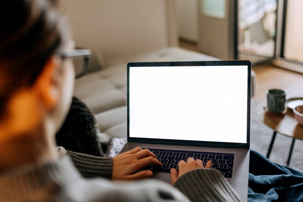 Over the shoulder view of young woman using laptop with white blank screen while relaxing on a sofa at home