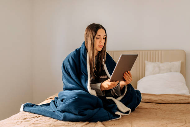 istock Beautiful young woman using digital tablet while sitting on a bed in bedroom wrapped in blanket 2198412986