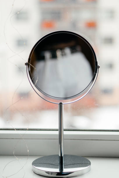 istock A round vanity mirror with a sleek chrome design stands on a windowsill, reflecting a blurred urban cityscape in the background, creating a modern and minimalistic aesthetic. 2198418340