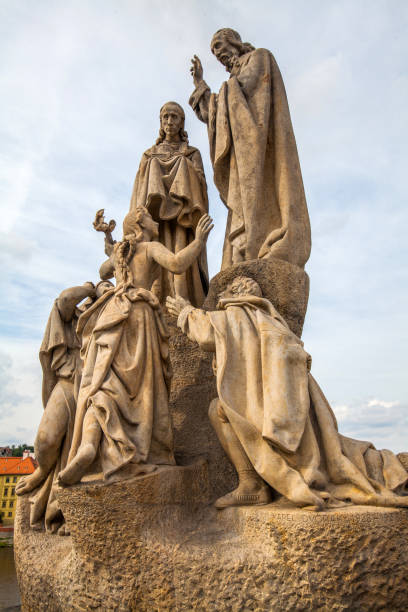 istock A statue group featuring several figures with detailed clothing, positioned atop a stone pedestal against a backdrop of a cityscape with red-roofed buildings and greenery under a cloudy sky. 2198426765