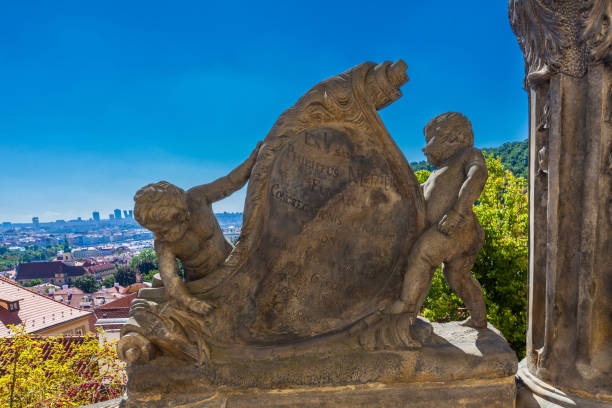 istock A stone sculpture featuring two cherubs holding a decorated shield with Latin inscriptions stands on a balcony overlooking a sprawling cityscape under a bright blue sky. 2198427845