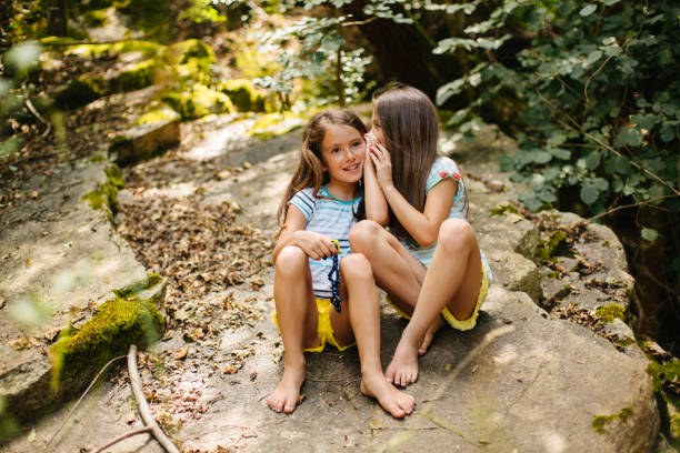 Two young girls enjoy a playful moment sitting outdoors in a tranquil forest environment. They are bonding and sharing secrets, immersed in nature, enjoying a beautiful, relaxed, and carefree day together.