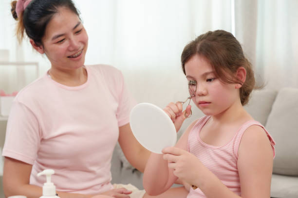 istock Caucasian girl learning makeup with Asian mom. Little girl using eyelash curler while looking into mirror. Mother smiling warmly, watching in cozy home setting with beauty products on table. 2198441419
