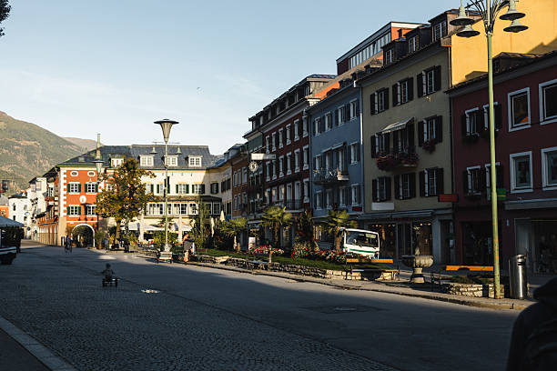A charming street in an Alpine town during the golden hour, featuring colorful buildings with traditional European architecture. The cobblestone street and flower-decorated façades create a welcoming atmosphere, while a cyclist and a child playing add a touch of life to the tranquil scene. Mountains in the background complete the idyllic setting.