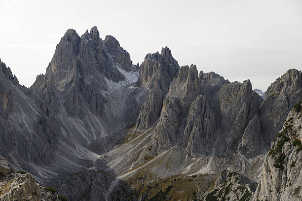 A dramatic view of the rugged peaks of the Dolomites in northern Italy. The towering rock formations, with their sharp ridges and steep slopes, dominate the landscape. Patches of greenery contrast with the gray limestone, highlighting the raw and untamed beauty of this alpine region.