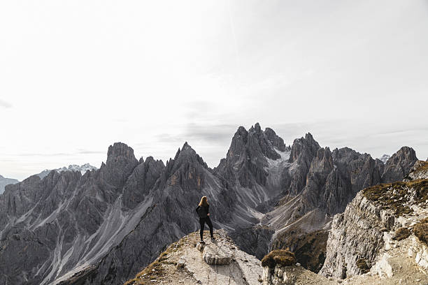 Female hiker standing on a rocky trail, overlooking the dramatic jagged peaks of the Dolomites mountain range in Italy. The solitary figure emphasizes the scale and ruggedness of the alpine landscape under a cloudy sky.