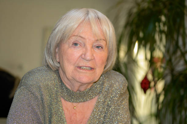 An elderly woman with silver hair and bright blue eyes sits in an indoor setting, dressed in a shimmering silver outfit with a stylish neckline.
