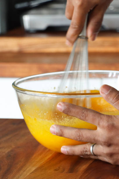 istock Full frame image of glass mixing bowl on wooden kitchen counter, unrecognisable person using stainless steel balloon whisk to mix eggs and sugar, beaten egg, wooden counter in kitchen setting, focus on foreground 2198481850