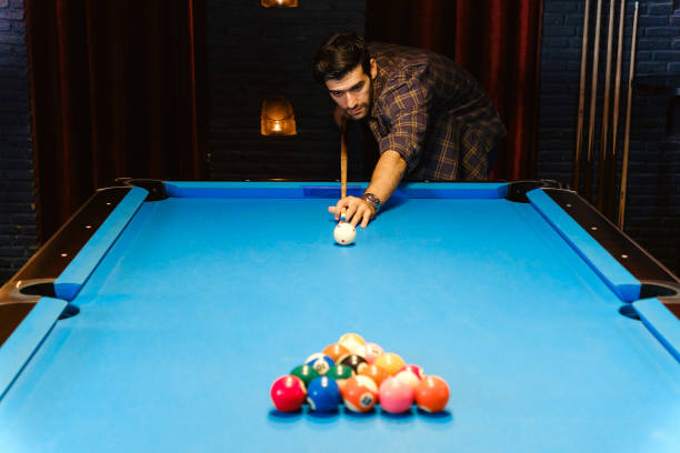 istock Focused man lines up his shot while playing billiards in a dimly lit pool hall. Blue pool table and colorful billiard balls create a competitive and stylish atmosphere, representing leisure, precision 2198484788