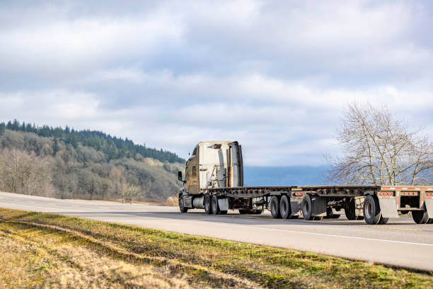 istock Beige big rig high cab semi truck with two empty flat bed semi trailers running on the road with forest on the hillside 2198488427