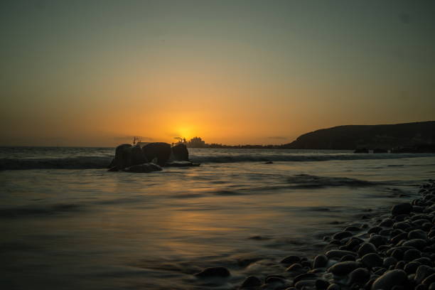 Rocky beach in Gran Canaria at sunset, sunrise. Smooth, rounded rocks in the foreground with a warm, glowing sky over the ocean. Tranquil coastal scene.