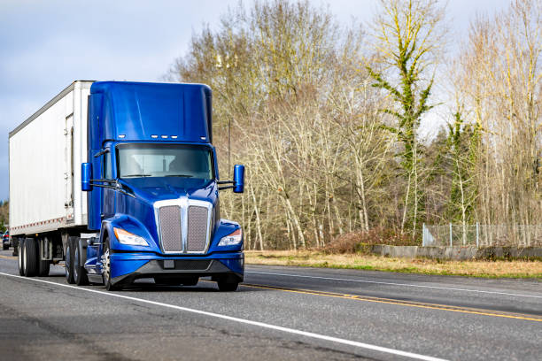 istock Bright blue big rig day cab semi truck transporting cargo in refrigerator semi trailer running on the highway road with bare winter trees on the side 2198488982
