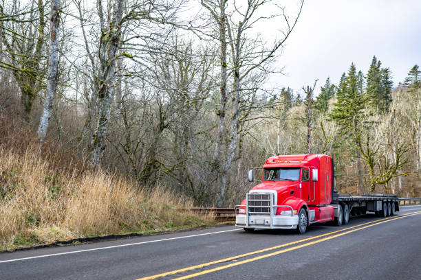 istock Red bonnet big rig semi truck with extended cab and grille guard transporting empty flat bed semi trailer driving on the narrow highway road with winter forest 2198489822