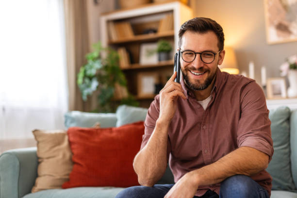 istock Young man is sitting on a sofa at home, engaged in a phone conversation. The scene captures a moment of communication, relaxation, or multitasking in a comfortable living space. 2198490467