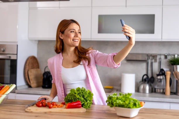istock A woman in a modern kitchen prepares dinner while taking a selfie, capturing a fun and joyful moment. She is multitasking, cooking a delicious homemade meal while enjoying the process. 2198490894