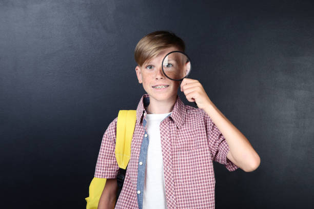 istock Young boy with magnifying glass and backpack on blackboard background 2198508778
