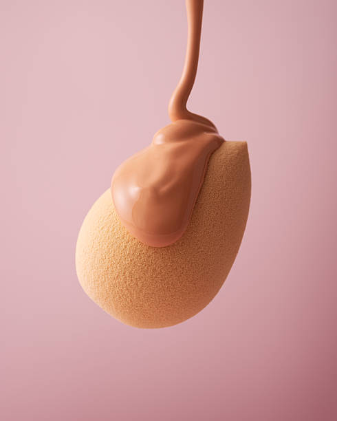 A close-up of a makeup sponge blender with liquid foundation being poured over it against a soft pink background