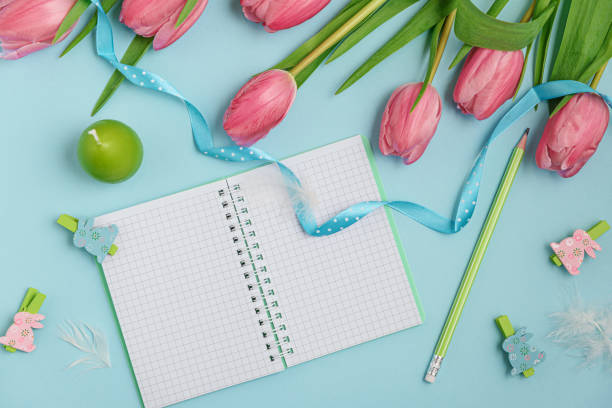 A pastel Easter scene featuring a bouquet of pink tulips adorned with a delicate blue ribbon, a notebook, wooden rabbit figurines, and soft feathers. Perfect for celebrating the Easter holiday.