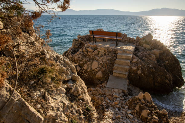 A serene coastal view of a cozy bench placed on rocky terrain, offering a perfect spot to relax while gazing at the shimmering water and surrounding mountains.