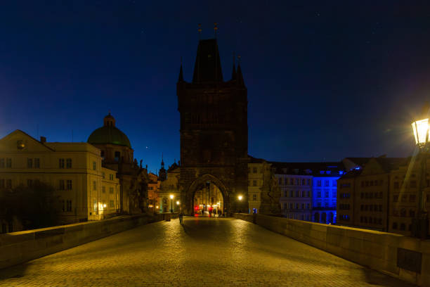 istock A stone bridge leading towards a tall tower at night, illuminated by streetlights, with a cityscape glow and a dark blue sky above. 2198548159
