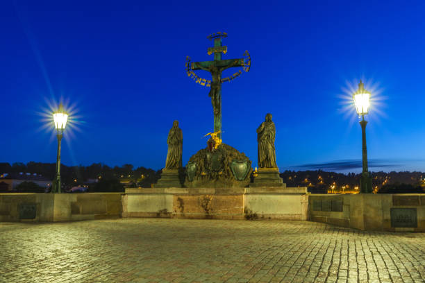 A stone bridge at twilight featuring a central religious monument with a crucifix and statues, illuminated by streetlights, with a cityscape in the background under a deep blue sky., Charles Bridge