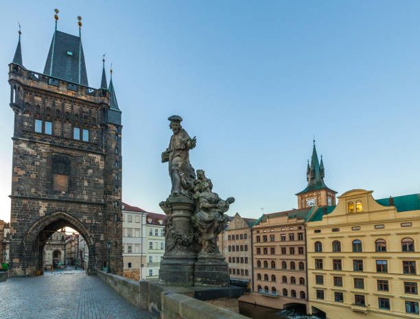 istock A stone bridge with a statue and a tall, dark tower with spires, leading into a cityscape with buildings and a clock tower under a clear sky at dusk. 2198553151