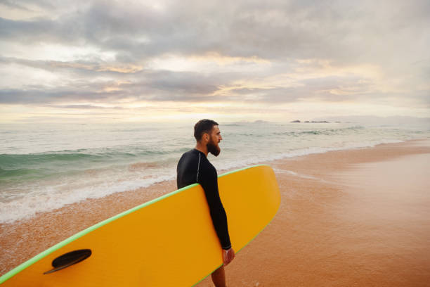 istock Young handsome male surfer with a surfboard is enjoying a view while walking a sandy beach at sea. Summer, vacation, ocean 2198555354