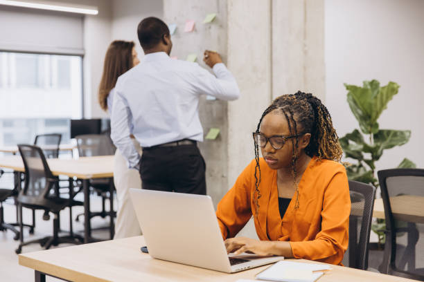 istock Black female entrepreneur working on laptop, focusing intently while team members brainstorming nearby in modern startup workspace 2198564163