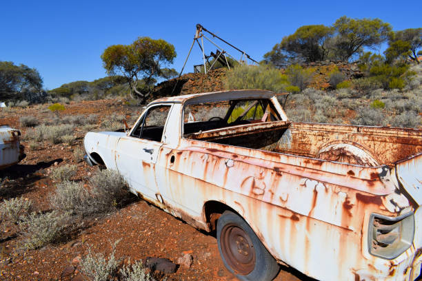 A white car left to decay in the vast desert near Kalgoorlie, Western Australia, with an old abandoned gold mine in the background.