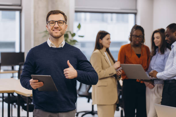 istock Confident businessman displaying tablet, gesturing approval with thumbs up while colleagues collaborating in contemporary workspace 2198564586