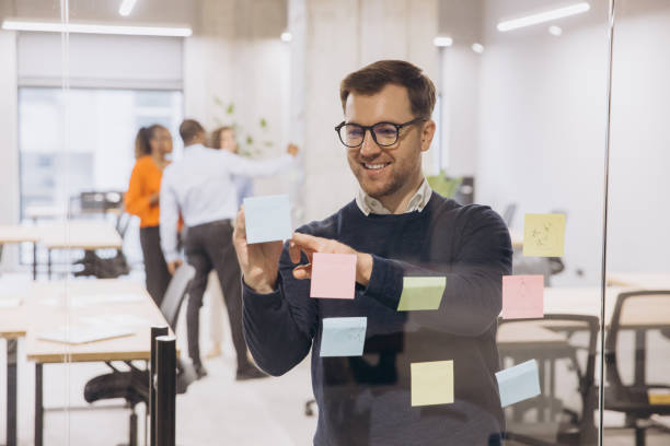 istock Smiling businessman using sticky notes on glass wall in a modern office, collaborating with colleagues in the background for a team project 2198568348