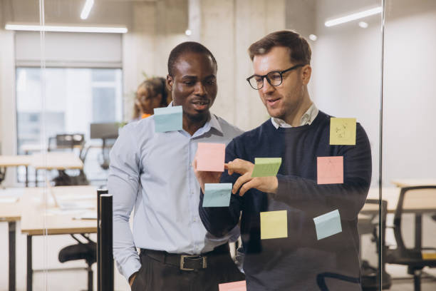 istock Two businessmen discussing ideas with sticky notes on a glass wall in a modern office, showcasing teamwork and collaboration 2198568350