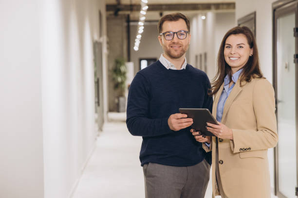 istock Smiling business colleagues holding a digital tablet while standing in a modern office hallway, engaging in productive discussion and collaboration 2198571068