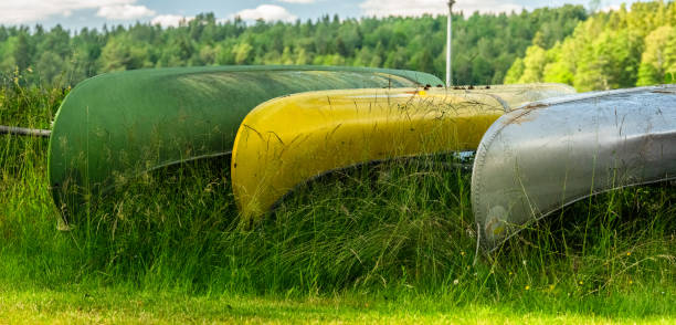 Three colorful boats turned upside down and stored by a fence on a grassy roadside. Seasonal outdoor recreation, infrastructure for water activities, and rental services in rural areas. Suitable for eco-tourism, summer travel, sports equipment, lakeside life