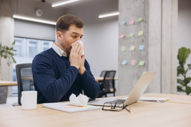 istock Businessman sneezing into a tissue while working at his desk, struggling with cold symptoms and feeling unwell in the office environment 2198573540