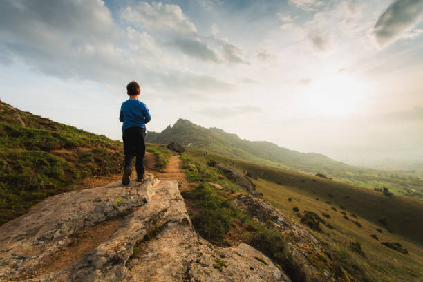 A young boy walking on a rocky mountain. The background features a vast green valley with mountain range. The sky is partly cloudy, with soft sunlight creating a peaceful atmosphere. The boy is waking on a rural path and looking at the sunset.
