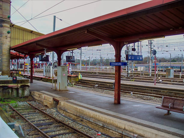 A view of a train platform, featuring a red-brown covered waiting area, multiple train tracks, and a sign indicating \
