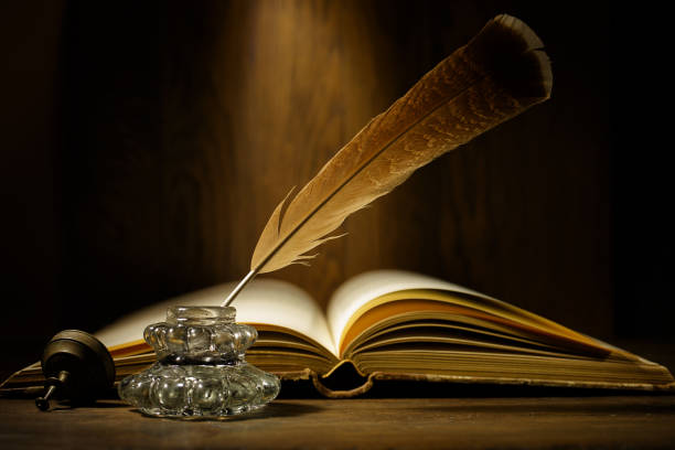 istock Open antique book with feather and ink bottle on old wooden table. Feather for calligraphy, old education, vintage still life. 2198586797