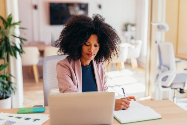 istock Focused young businesswoman taking notes while working on a laptop in a modern office 2198589595