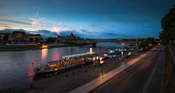 Colorful fireworks illuminate the night sky over the Elbe River. Boats are docked along the riverbank as people enjoy the festivities, creating a vibrant atmosphere in Dresden.