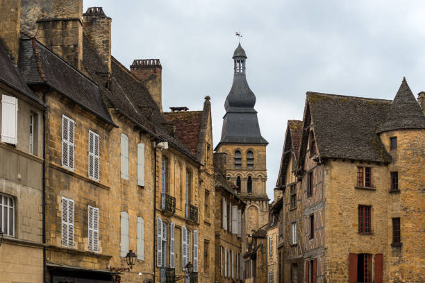 Visitors stroll through the charming streets of Sarlat-la-Caneda, surrounded by historic stone buildings. The iconic tower stands tall amidst the overcast sky, creating a picturesque setting.