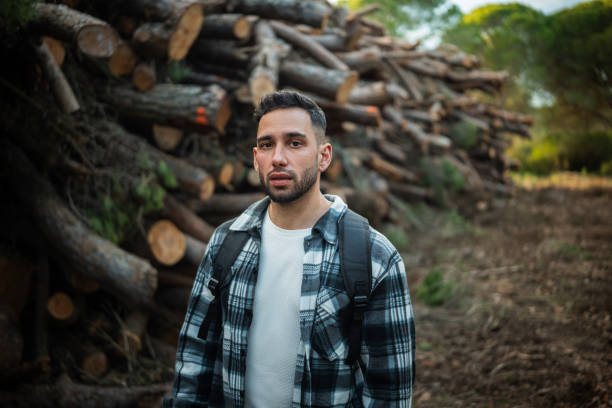Young lumberjack with backpack posing near a pile of logs in a forest