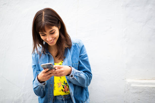 istock Smiling young woman leaning against white wall and using cellphone 2198602923