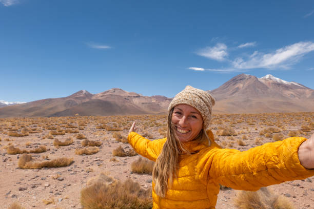 Smiling woman in yellow jacket taking selfie in arid desert with mountains. Adventurous traveler in vast wilderness. Capturing nature's beauty in remote landscape.