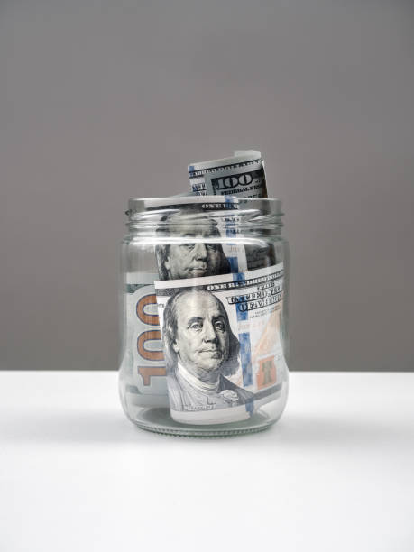istock One hundred us dollars bill banknotes cash with Benjamin Franklin's face in glass jar isolated on white table and grey background, vertical style. Saving money, budget, tax, financial concept. 2198617640