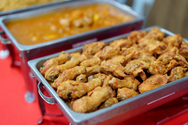 A tray filled with crispy golden-brown fried chicken drumettes and wings, served at a buffet-style catering event. A container of curry dish is seen in the background.