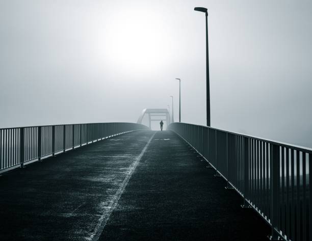 A lone figure walks on a foggy bridge, with street lamps and railings, creating a mysterious atmosphere.