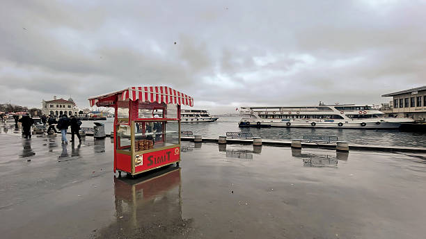 Kadikoy,Istanbul,Turkey.January 12,2025.Kadıköy Coastal. Rainy Day Flavors on the Bosphorus. A lone red and white striped bagel stand stands on a wet, reflective sidewalk near the Bosphorus Sea in Istanbul. The cloudy sky and blurred figures in the background evoke a rainy day. In the distance, ferries and waterfront buildings can be seen through the foggy atmosphere. The stand, presumably serving snacks or drinks, is closed, its vibrant colors contrasting with the pale tones of the surrounding environment.